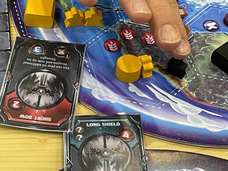 Et brætspil med kort, spilleplader og brikker - og en hånd der flytter på en brik på en af spillepladerne